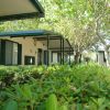 Esterno di bungalow con tettoia, finestre verdi e piante rigogliose attorno.