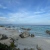 Spiaggia rocciosa con sabbia bianca e mare azzurro sotto un cielo nuvoloso.