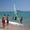 Ragazzi sulla spiaggia osservano un windsurfista nel mare blu con piccola isola sullo sfondo.