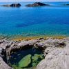 Laghetto roccioso vicino a un mare azzurro con isole in lontananza.