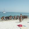 Persone giocano al tiro alla fune su una spiaggia di fronte al mare con barche a vela in lontananza.