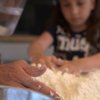 Mani che impastano farina, con una bambina sullo sfondo che osserva.