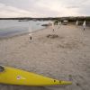Spiaggia sabbiosa con kayak giallo e ombrelloni chiusi.