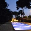 Piscina illuminata di notte con luci blu, circondata da alberi e ombrelloni.