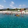 Mare limpido con persone che nuotano verso la spiaggia, ombrelloni verdi e montagne sullo sfondo.