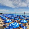 Spiaggia con file di ombrelloni blu e lettini, bandiere sventolano in lontananza.