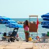 Spiaggia con ombrelloni blu, persone sedute, passeggino e mare sullo sfondo.