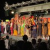 Un gruppo di attori saluta il pubblico su un palco, indossando costumi e maschere colorate.