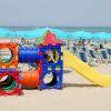 Gioco colorato per bambini su una spiaggia con sedie a sdraio blu e ombrelloni sullo sfondo.