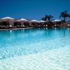 Piscina all'aperto con lettini e ombrelloni bianchi, cielo limpido e palme sullo sfondo.