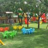Area giochi con tavolini colorati, scivoli e giostre tra gli alberi.