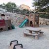 Parco giochi con scivoli, altalene e tavolo da picnic su sabbia.