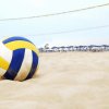 Palla da volley su una spiaggia sabbiosa con rete e ombrelloni blu sullo sfondo.