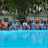 Persone in piscina che spruzzano acqua, sfondo con alberi.