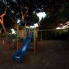 Scivolo blu e altalene in un parco giochi ombreggiato.
