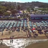 Vista aerea di una spiaggia affollata con ombrelloni azzurri e un chiosco.
