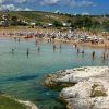 Spiaggia con ombrelloni blu e bianchi, persone in acqua limpida, rocce in primo piano.