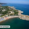 Panorama costiero con spiaggia affollata e mare blu, vegetazione e edifici lungo la costa.