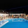 Una piscina illuminata di notte con persone sedute e in piedi intorno, sotto luci decorative.