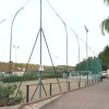 Campo sportivo recintato con luci, piante e una passerella pavimentata.
