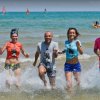 Cinque persone corrono sorridenti in mare, con vele sullo sfondo.