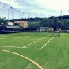 Campo da tennis all'aperto con rete e righe bianche, circondato da reti alte e vegetazione.