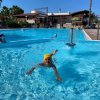 Bambino che nuota in una piscina all'aperto con giochi d'acqua e sfondo di edifici e palme.