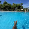 Gambe di una persona immerse in una piscina limpida, circondata da palme e vegetazione.