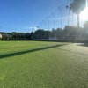 Campo da calcio in erba sintetica con rete di protezione e luci, illuminato dal sole.