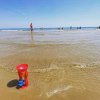 Giocattolo rosso in riva al mare con persone che camminano nell'acqua bassa e cielo azzurro.