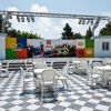 Piazzale con tavoli, sedie e palco colorato.