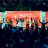 Un gruppo di persone balla su un palco illuminato, con uno sfondo colorato e un pubblico davanti.