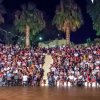 Folla seduta su gradinate all'aperto di notte, con palme e festoni colorati.