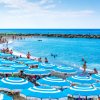 Spiaggia con molti ombrelloni blu e bianchi, mare calmo e persone che nuotano.