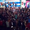 Persone in piedi, ballano sotto un palco illuminato. Sul palco, un gruppo di ballerini si esibisce.
