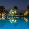 Una piscina illuminata di sera con palme e alberi riflessi nell'acqua.