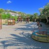 Parco giochi con scivoli, altalene e giostra circolare, contornato da palme.