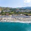 Spiaggia affollata con ombrelloni blu e bianchi, mare azzurro e colline in lontananza.