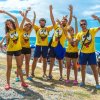 Gruppo di persone in magliette gialle con disegni sorridenti, sfondo mare e ombrelloni.