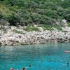 Costa rocciosa con acqua cristallina e persone che nuotano.