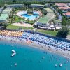 Lido affollato con ombrelloni gialli e blu, piscina circolare e edifici residenziali.