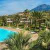Un'ampia piscina circondata da lettini, ombrelloni e palme, con montagne sullo sfondo.