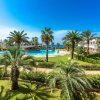 Piscina circondata da palme, giardino e costruzioni, mare visibile in distanza.