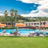 Piscina all'aperto con ombrelloni arancioni e lettini, circondata da palme e prati.