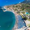 Spiaggia affollata con file di ombrelloni blu e mare calmo, vista su colline e case costiere.