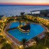 Piscina illuminata di notte, circondata da palme e edifici, con vista sul mare al tramonto.