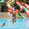 Bambina salta in piscina con braccioli. Scivolo colorato sullo sfondo.