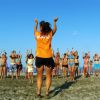 Gruppo di persone fa esercizi in spiaggia, guidati da una donna di spalle in maglietta arancione.