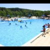 Piscina all'aperto affollata di persone, circondata da ombrelloni e vegetazione.