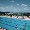 Piscina all'aperto con bagnanti e lettini, colline sullo sfondo.